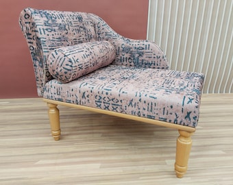 May include: A chaise lounge with a patterned fabric in shades of pink and teal. The chaise has a rounded back, a cylindrical bolster pillow, and light-colored wooden legs. The fabric features a geometric design.