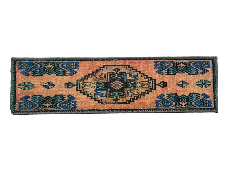 Puede incluir: Una alfombra larga y estrecha con un dise&ntilde;o turco tradicional. La alfombra tiene un fondo melocot&oacute;n con un patr&oacute;n central en forma de diamante en tonos de azul, verde y negro. El patr&oacute;n de diamante est&aacute; rodeado de formas geom&eacute;tricas m&aacute;s peque&ntilde;as en los mismos colores. La alfombra tiene un borde gris oscuro.