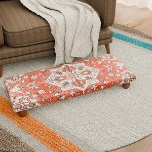 May include: A rectangular footstool with a red and white patterned design, featuring a traditional rug motif. The footstool has wooden legs and is placed on a light gray rug. A brown armchair and a cream-colored blanket are in the background.