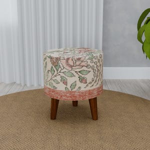 May include: A round, floral patterned ottoman with a red base and four wooden legs. The ottoman is sitting on a beige rug.
