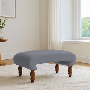 May include: A gray upholstered ottoman with four wooden legs. The ottoman has a curved design and is placed on a patterned rug. The legs are a dark brown color.