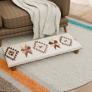Puede incluir: Un reposapiés rectangular con una parte superior de tela color crema con patrones geométricos en tonos marrones. El reposapiés tiene cuatro patas de madera y está colocado sobre una alfombra gris claro con rayas coloridas. Un sillón marrón y una manta color crema en el fondo.
