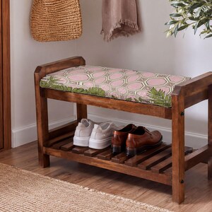 May include: A dark wood shoe bench featuring a rectangular seat cushion with a pink and green geometric and botanical pattern. The bench has a slatted lower shelf for storage, currently holding a pair of white sneakers and brown leather shoes.