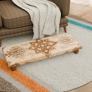 Puede incluir: Un reposapiés rectangular con un diseño beige y marrón, apoyado sobre cuatro patas de madera. El reposapiés está colocado sobre una alfombra gris claro, con un sillón marrón y una manta blanca en el fondo.