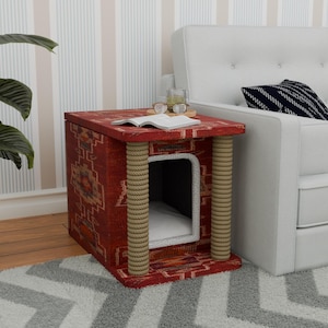Può includere: Un letto per gatti di colore rosso e crema con un bordo in corda. Il letto ha la forma di una casa e ha una piccola apertura per consentire al gatto di entrare. Il letto è posizionato su un tappeto bianco davanti a un divano bianco.