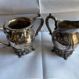 May include: A silver sugar bowl and creamer set with ornate detailing and a footed base.