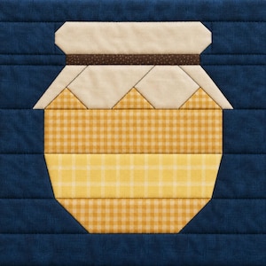 May include: A fabric quilt block featuring a central jar motif against a solid dark blue background. The jar is constructed from beige, yellow, and brown patterned fabric pieces, with a horizontal brown band separating the lid from the body.