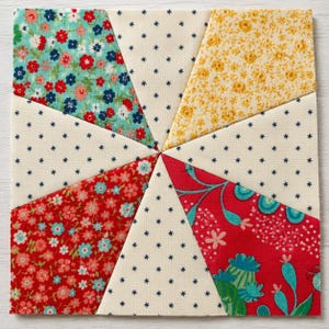 May include: A square quilt block featuring a fan-like arrangement of eight triangular fabric segments. The design alternates between cream fabric with small dark blue star patterns and various floral prints in shades of turquoise, red, and yellow.