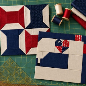May include: Two square fabric quilt blocks featuring geometric patterns in red, white, and navy blue. One block displays a pinwheel design, while the other depicts a sewing machine silhouette with a heart motif. Spools of thread and fabric scraps are nearby.