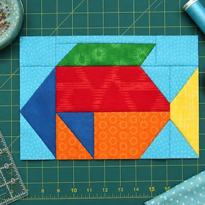 Può includere: Un blocco per trapuntatura rettangolare con un design a forma di pesce realizzato con vari ritagli di tessuto colorati. Il corpo del pesce è rosso, con una pinna superiore verde, una coda gialla e sezioni inferiori blu e arancioni su sfondo azzurro.