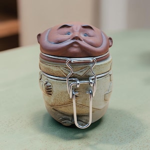 Może przedstawiać: Ceramiczny pojemnik z pokrywką w kształcie ludzkiej głowy z wąsami i brązowym odcieniem skóry. Podstawa jest w kolorze nakrapianego beżu, uformowana tak, aby przypominała tułów z ramionami i ubraniem. Srebrne metalowe zamknięcie zabezpiecza pokrywkę.