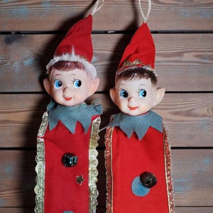 Puede incluir: Dos muñecos de duendes navideños vintage con trajes de fieltro rojo, gorros puntiagudos y caras sonrientes. Los muñecos tienen ojos azules, cuellos grises y adornos dorados. Uno tiene una gema verde y una estrella, el otro una campana.