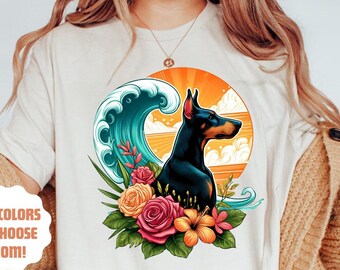 May include: A white t-shirt with a graphic of a Doberman Pinscher standing in front of a sunset with a wave and flowers. The text '+10 colors to choose from!' is on the left side of the shirt.