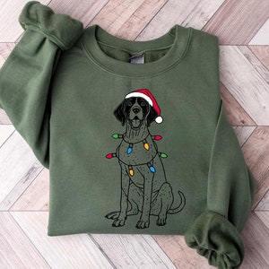 Puede incluir: Una sudadera verde con una ilustración en blanco y negro de un perro con un gorro de Papá Noel rojo y luces de Navidad.