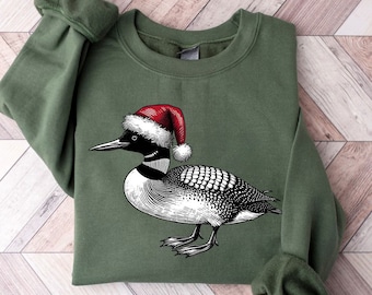 May include: An olive green sweatshirt featuring a detailed illustration of a loon wearing a red and white Santa hat. The loon is depicted in black and white, with intricate feather detailing. The sweatshirt has a crew neck and long sleeves.