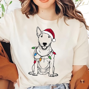 Könnte beinhalten: Weißes T-Shirt mit einer Schwarzweißillustration eines Bullterriers, der einen roten Weihnachtsmann-Hut und Weihnachtslichter trägt.
