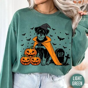 Puede incluir: Sudadera verde claro con un diseño de Halloween. El gráfico muestra un perro negro con sombrero de bruja y capa naranja, junto a tres calabazas talladas. Murciélagos y una lápida con "RIP" completan la escena espeluznante.