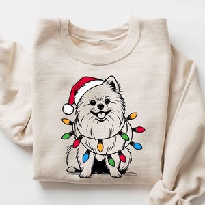 May include: A beige sweatshirt featuring a graphic of a white dog wearing a red Santa hat and a string of colourful Christmas lights around its neck.