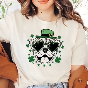 May include: White t-shirt with a cartoon pug wearing a green leprechaun hat and heart-shaped sunglasses. The pug is surrounded by green shamrocks.