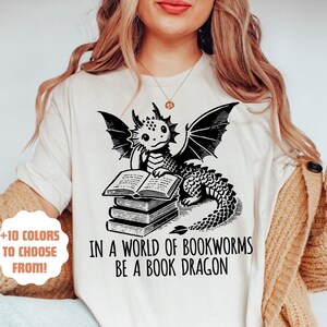 May include: A white t-shirt featuring a black illustration of a dragon reading a book. The text on the shirt reads "In a world of bookworms be a book dragon".