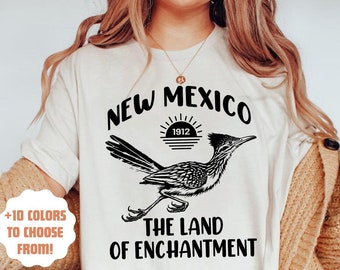 Könnte beinhalten: Ein weißes T-Shirt mit einem schwarzen Grafikdesign, das einen Roadrunner, einen Sonnenuntergang und den Text "New Mexico 1912 The Land of Enchantment" zeigt.