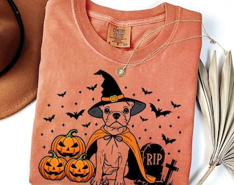 Puede incluir: Una camiseta de color melocotón con una ilustración en negro y naranja de un perro con un sombrero de bruja y una capa. El perro está rodeado de calabazas y murciélagos. El texto "RIP" está en una lápida en la imagen.