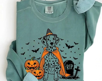 May include: A teal blue sweatshirt with a Halloween-themed design featuring a Dalmatian dog wearing a witch's hat and a cape, holding a pumpkin. The dog is standing in front of a tombstone that says "RIP" and there are pumpkins and bats in the background.