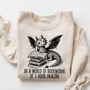 May include: A beige sweatshirt with a black graphic of a dragon reading books. The dragon is sitting on a stack of books. The text "IN A WORLD OF BOOKWORMS BE A BOOK DRAGON" is printed below the dragon.