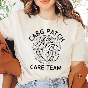 Puede incluir: Camiseta color crema con el texto negro "CABG PATCH CARE TEAM" y un gráfico de un corazón dentro de una col. La camiseta está hecha de un material suave.