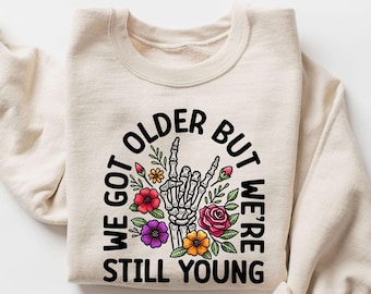 May include: A beige sweatshirt with a black graphic print that says "We got older but we're still young". The graphic features a skeleton hand giving a rock and roll sign with flowers around it.