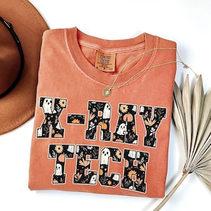 Puede incluir: Una camiseta naranja quemada con un gráfico negro que dice "X-RAY TECH" con fantasmas y calabazas en las letras.