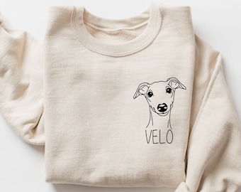 Könnte beinhalten: Ein beiger Pullover mit einer schwarzen Strichzeichnung eines Hundekopfes und dem Namen "VELO" auf der Vorderseite.