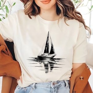 以下が含まれることがあります： 白と黒のヨットのグラフィックが特徴のクリーム色のTシャツ。デザインは、水面に映るヨットを描いています。シャツはクラシックなクルーネックと半袖です。全体的なスタイルはカジュアルで航海的です。
