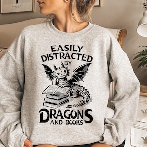 May include: A gray sweatshirt with a black graphic of a dragon reading a book. The text on the sweatshirt reads "Easily Distracted by Dragons and Books."