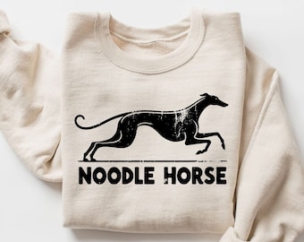 Pode incluir: Camisola creme com um gráfico preto de um cão a correr e as palavras "NOODLE HORSE" em negrito e com aspecto envelhecido. A camisola tem mangas compridas e gola redonda.
