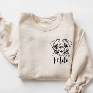 May include: A cream-coloured sweatshirt with a black outline of a pug's face and the name "Milo" beneath. The sleeves are rolled up, giving a relaxed look. The design is simple and suitable for pet lovers. The sweatshirt is folded.