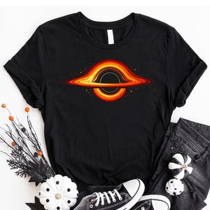 May include: Black t-shirt featuring a colorful graphic of a black hole with orange, yellow, and red hues. The shirt is paired with black jeans and black and white sneakers. Decorative pumpkins and branches complete the ensemble.