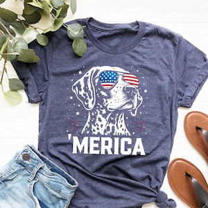 May include: Navy blue t-shirt featuring a dog wearing sunglasses with an American flag design. The word "'MERICA" is printed below the dog. The shirt is paired with denim shorts and sandals.
