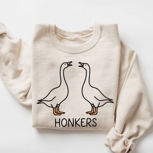 Könnte beinhalten: Ein beiger Pullover mit einer Grafik von zwei Gänsen, die sich gegenüberstehen, und dem darunter gedruckten Text "HONKERS".