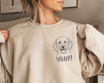 May include: A beige sweatshirt with a black line drawing of a dog on the front. The dog is smiling and the text "sunny" is written below the dog.
