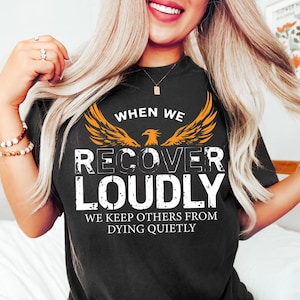 Könnte beinhalten: Schwarzes T-Shirt mit einem orangefarbenen Adler-Motiv und dem Text "When we recover loudly we keep others from dying quietly."