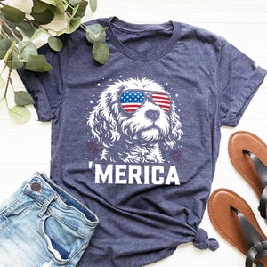May include: Navy blue t-shirt with a white dog wearing sunglasses in the style of the American flag. The word "'MERICA" is printed below. The image includes denim shorts and brown sandals.