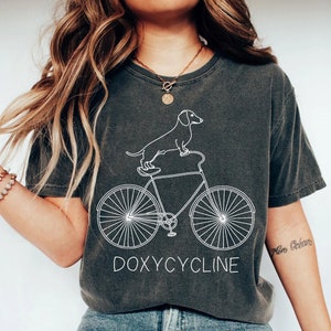 May include: A dark gray t-shirt with a white outline of a dachshund riding a bicycle. The text "DOXYCYCLINE" is printed below the image.