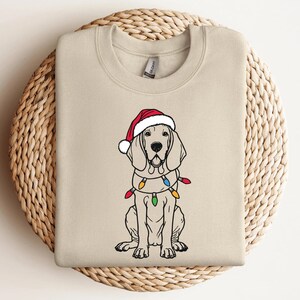 May include: A beige sweatshirt with a Christmas-themed illustration of a dog wearing a Santa hat and a string of colorful lights around its neck.