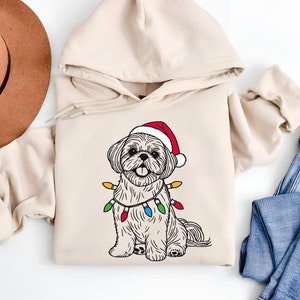 Op de afbeelding: Een beige hoodie met een kerstthema-illustratie van een Shih Tzu-hond die een kerstmanmuts draagt en een reeks kleurrijke lampjes om zijn nek heeft.