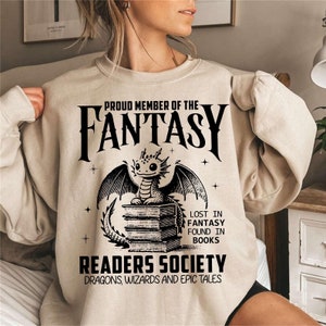 May include: A beige sweatshirt with a black graphic print of a dragon sitting on a stack of books. The text reads "Proud Member of the Fantasy Readers Society Dragons, Wizards and Epic Tales".