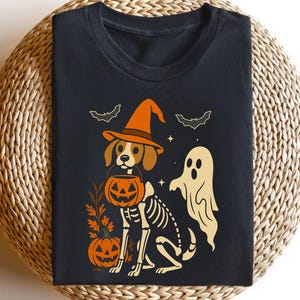 May include: Black t-shirt with a Halloween-themed design. The design features a dog in a witch's hat, holding a pumpkin lantern, with a skeleton body. A ghost and bats are also included. The t-shirt is folded on a woven surface.