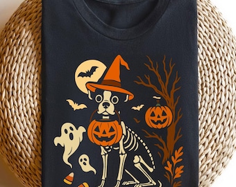 Puede incluir: Camiseta negra con un diseño de Halloween. Presenta un esqueleto de perro con un sombrero de bruja y una linterna de calabaza. El diseño también incluye fantasmas, murciélagos, una luna llena y una calabaza.