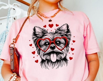 Op de afbeelding: Een roze T-shirt met een zwart-witte illustratie van een Yorkshire Terrier die een rode hartvormige bril draagt. De hond is omringd door kleine rode hartjes.