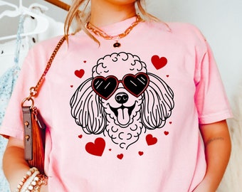 May include: A pink t-shirt with a black and white illustration of a poodle wearing heart-shaped sunglasses. The poodle is surrounded by red hearts.